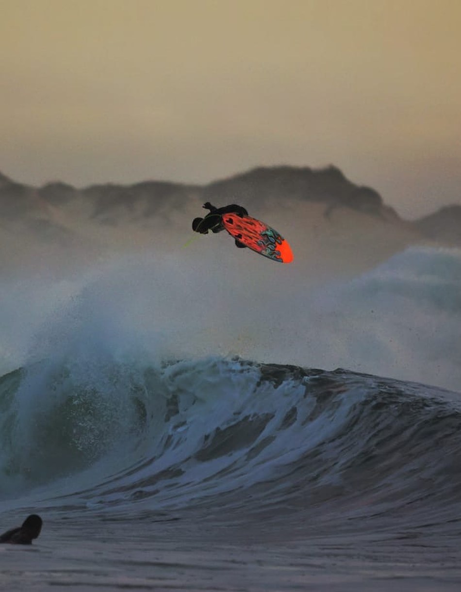 A surfer launching an air above a breaking wave at dusk, dunes behind
